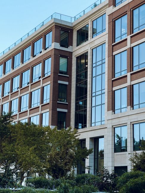 Exterior view of a modern multi-storey commercial office building with large glass windows and brick façade, reflected in the glass panels. The building features a combination of brown brick and light-colored stone accents. In the foreground, lush green trees and shrubs are visible, enhancing the landscaped environment. The clean and well-maintained appearance of the building suggests regular cleaning and upkeep by professionals such as those from Cleaner Holland Park, who specialize in surface cleaning, deep cleaning, and sanitisation for commercial properties. Natural daylight illuminates the scene, highlighting the building's polished glass surfaces and tidy exterior. This setting exemplifies professional property maintenance aimed at maintaining hygiene and a professional aesthetic for business premises in Holland Park and Kensington. 