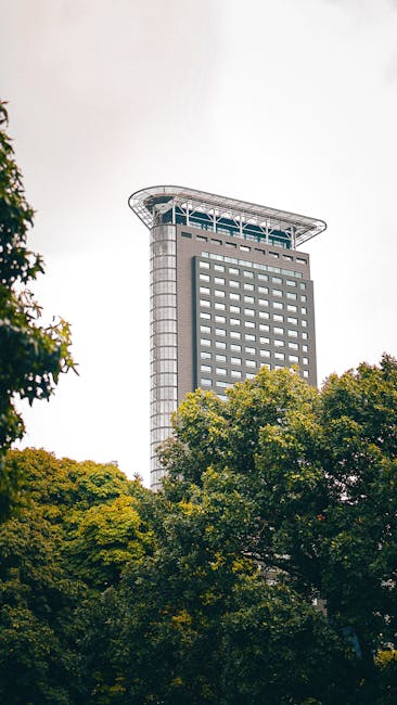 The image showcases a tall, modern office building with a distinctive curved rooftop structure, constructed with glass and steel materials, situated behind a cluster of lush green trees. The building's reflective glass windows display a sleek, clean appearance, indicating recent surface cleaning and maintenance. The overcast sky provides diffuse lighting that highlights the building's architectural features and the vibrant greenery in the foreground. This setting emphasizes the importance of professional cleaning and upkeep for commercial properties like this, aligning with the services offered by Cleaner Holland Park, who provide comprehensive surface cleaning, deep cleaning, and sanitisation for business environments in Holland Park and Kensington.