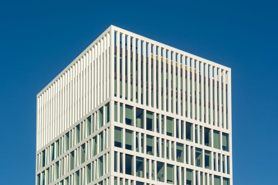 Exterior view of a modern multi-storey commercial office building with large glass windows and brick façade, reflected in the glass panels. The building features a combination of brown brick and light-colored stone accents. In the foreground, lush green trees and shrubs are visible, enhancing the landscaped environment. The clean and well-maintained appearance of the building suggests regular cleaning and upkeep by professionals such as those from Cleaner Holland Park, who specialize in surface cleaning, deep cleaning, and sanitisation for commercial properties. Natural daylight illuminates the scene, highlighting the building's polished glass surfaces and tidy exterior. This setting exemplifies professional property maintenance aimed at maintaining hygiene and a professional aesthetic for business premises in Holland Park and Kensington. 