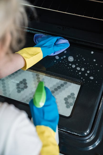 A person wearing blue and yellow rubber gloves is cleaning the glass and exterior of an oven with a cloth, applying surface cleaning and sanitisation to a black oven door. The oven has a ceramic or metal finish, with visible soap suds on the surface, and is situated in a kitchen setting. The internal reflection of baking trays or items can be seen through the oven glass, emphasizing a thorough deep cleaning process. Available lighting highlights the shiny, clean surface, typical of professional domestic cleaning services provided by Cleaner Holland Park, as seen on the website cleanerhollandpark.co.uk.