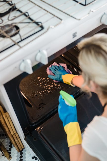 A professional cleaning technician from Cleaner Holland Park is performing deep oven cleaning in a domestic kitchen. The technician is wearing protective blue and yellow rubber gloves and is using a pink cloth to carefully wipe the black oven door, which shows a few splashes of soap or cleaning solution. The oven door, made of glass and surrounded by a white frame, is partially open, revealing the inside of the oven. Above the oven, a white gas stove with visible metal burners and control knobs is situated on a wooden countertop. The kitchen features a black and white patterned tile floor, and natural light illuminates the scene, emphasizing the cleanliness and attention to detail typical of thorough surface cleaning and sanitisation provided by Cleaner Holland Park, which specializes in deep cleaning services for both residential and commercial properties.