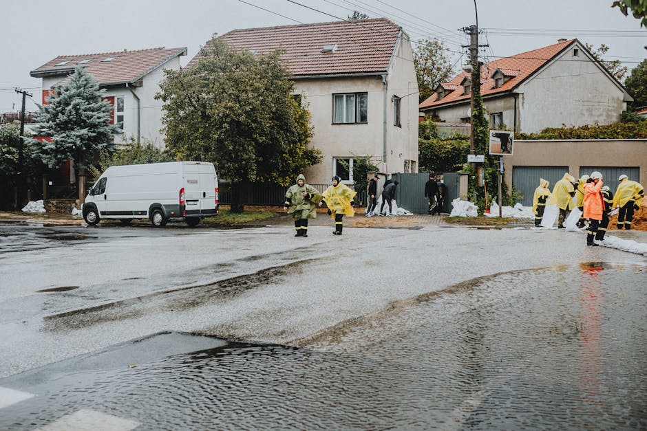 A residential street after flooding showing wet asphalt and water puddles, with a group of emergency responders and volunteers in yellow raincoats and protective gear working near the sidewalk and house front. A white van is parked on the side, and the background features multi-story houses with tiled roofs, one surrounded by trees and shrubs. The scene is set on an overcast day, emphasizing the need for surface cleaning and sanitisation services offered by Cleaner Holland Park to restore hygiene and tidiness after flood damage.