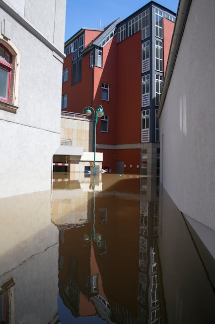 Image depicting an outdoor urban area affected by flooding, with muddy water covering parts of the pavement and street furniture. Tall modern buildings with red and grey facades surround the scene, and a streetlamp stands amidst the submerged area. The scene highlights the need for professional emergency flood cleaning, which Cleaner Holland Park provides through surface cleaning and sanitisation services to restore hygiene and safety to affected residential and commercial properties.