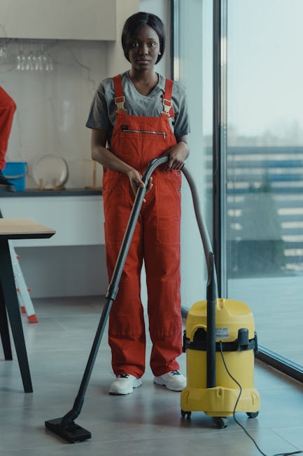 A professional cleaner from Cleaner Holland Park, dressed in red overalls and a grey t-shirt, is vacuuming a modern, light-colored tiled floor in a residential kitchen. The cleaner holds the vacuum hose firmly, guiding it over the surface to remove dust and debris, with the vacuum unit positioned beside her. The kitchen features a sleek, minimalistic design with a glass door leading to an outdoor balcony, allowing natural light to illuminate the space. Surfaces such as the kitchen counter and table are clear and tidy, emphasizing cleanliness. The setting showcases a well-maintained environment suitable for comprehensive surface cleaning services, reflecting the company's focus on deep cleaning and hygiene in domestic spaces.