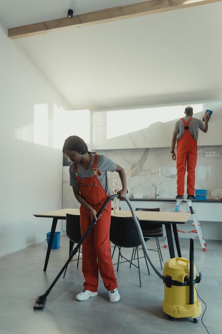 A professional cleaner from Cleaner Holland Park, dressed in red overalls and a grey t-shirt, is vacuuming a modern, light-colored tiled floor in a residential kitchen. The cleaner holds the vacuum hose firmly, guiding it over the surface to remove dust and debris, with the vacuum unit positioned beside her. The kitchen features a sleek, minimalistic design with a glass door leading to an outdoor balcony, allowing natural light to illuminate the space. Surfaces such as the kitchen counter and table are clear and tidy, emphasizing cleanliness. The setting showcases a well-maintained environment suitable for comprehensive surface cleaning services, reflecting the company's focus on deep cleaning and hygiene in domestic spaces.