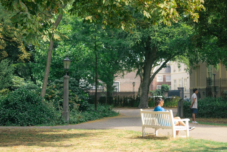 A woman seated on a white wooden bench in a residential park, engaging in surface cleaning or relaxation, with lush green trees and bushes providing shade. The area is illuminated by natural daylight, highlighting the well-maintained paths and green grass. In the background, a child stands nearby, while another person walks along the path. The scene emphasizes a clean, hygienic outdoor space maintained by professional cleaning services, reflecting the standards of Cleaner Holland Park for domestic and outdoor sanitisation, as seen on the Holland Park domestic cleaning services page. Surfaces and surroundings appear tidy and fresh, with no visible dirt or debris, exemplifying effective outdoor cleaning and maintenance practices.