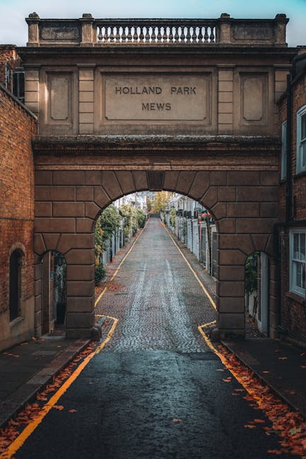 The image shows the entrance to Holland Park Mews, an arched brick gateway with a decorative stone balustrade on top, inscribed with 'Holland Park Mews.' The cobblestone street extends through the arch, flanked by residential buildings with brick facades and white-framed windows. The street is wet, possibly from recent cleaning or rain, with scattered fallen leaves along the edges. The scene is softly lit, highlighting the textures of the wet surface and brickwork. This setting exemplifies the tidy, well-maintained appearance typical of London’s residential mews, aligning with the high standards of clean surface maintenance and street hygiene associated with professional domestic cleaning services provided by Cleaner Holland Park.