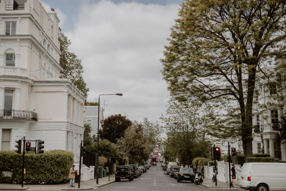 A row of multi-story residential buildings with varied architectural styles, including gabled roofs and large windows, are situated behind a lush landscape with mature trees and shrubs. In the foregro
